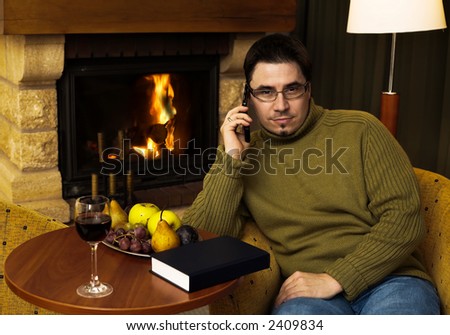 Man is sitting in the armchair at home in front of the fireplace. He talks on a cell phone and looks at the camera.