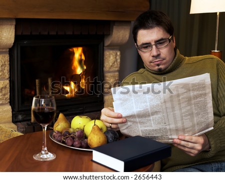 Man is sitting in the armchair at home in front of the fireplace. He is reading the newspaper.