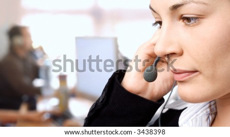 Young female customer service representative receives calls on a headset while an IT specialist works on a computer in the background.