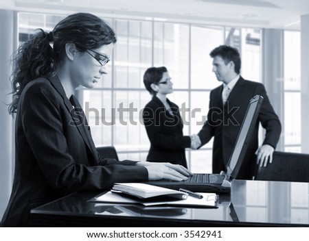 Young female secretary makes notes on a laptop  while other business people are shaking hands in the background. Daylight, indoor, office.