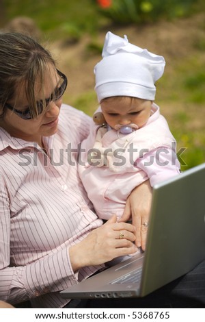Mother of a 9 months old baby-girl is sitting on the ground in the garden and uses a laptop computer.