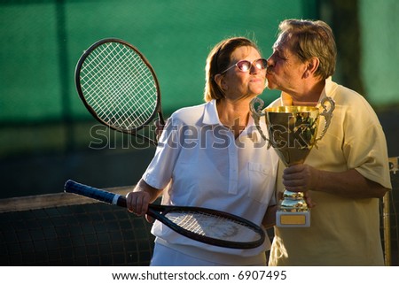 Active senior couple is posing on the tennis court with tennis racket and cup in hand. Outdoor, sunlight.