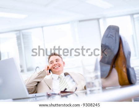 Satisfied businessman sitting by desk at office and calling on phone, feet on table, smiling.