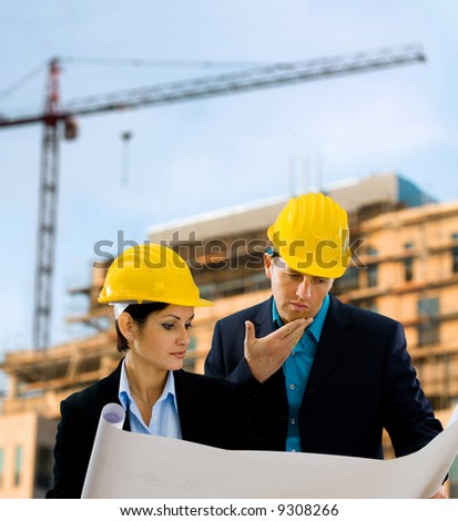 Young architects looking at blueprint in front of construction site, building and crane.