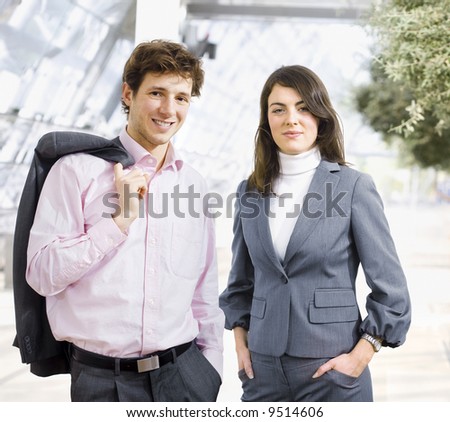 Happy young businessman and businesswoman standing side by side inside office building, smiling, looking at camera.