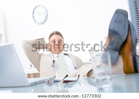 Satisfied businessman sitting by desk at office, feet on table, smiling, looking at camera.