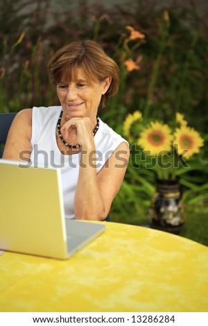 Modern senior woman sitting in the garden at home and using a laptop computer. Summer, outdoor.