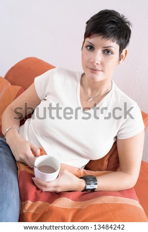 Young woman sits on the sofa at home in a relaxing pose and she drinks coffee. She is looking at the camera.