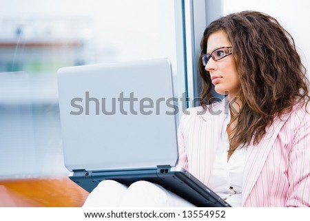 Young businesswoman sitting at office window using laptop computer and thinking.