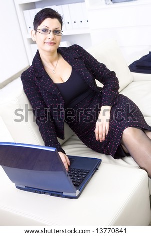 Happy businesswoman sitting on sofa at office and working on laptop computer, looking at camera, smiling.