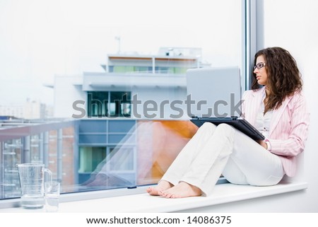 Young creative businesswoman sitting at office window using laptop computer and thinking.