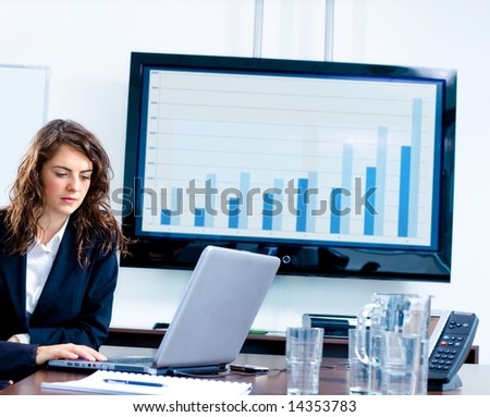 Young businesswoman sitting by meeting table at office in front of a huge plasma TV screen and using laptop and phone.