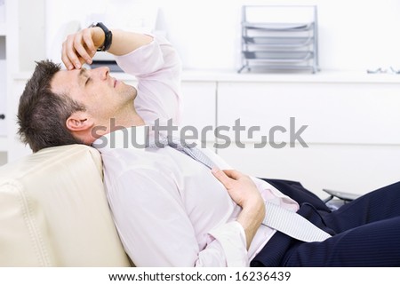 Mid-adult businessman lying on couch at office, looking exhausted eyes closed. Bright background.