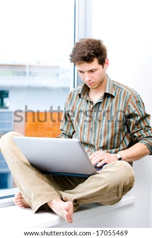 Young casual office worker sitting at office window working on laptop computer.