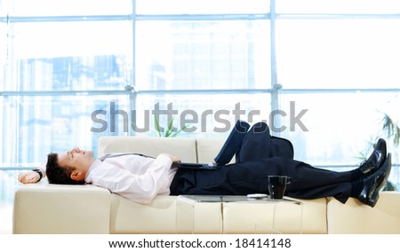 Businessman taking break at office, lying on back and thinking while resting on sofa and holding laptop computer.