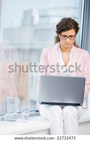Young attractive female office worker working on laptop computer in fron front of office window.