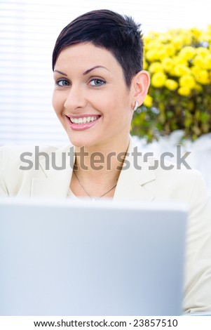 Businesswoman working on laptop computer in brightly lit office, smiling, looking at camera.