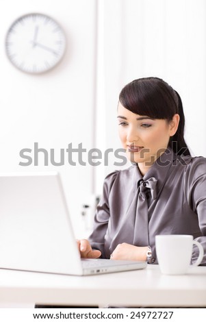 Businesswoman working on laptop in brightly lit office, smiling.