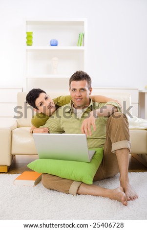 Young couple using laptop computer at home, sitting on floor and lying on couch, embracing.
