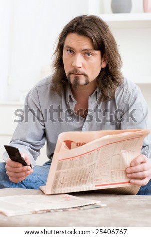 Man using mobile phone and holding newspaper in hand, sitting in living room at home.