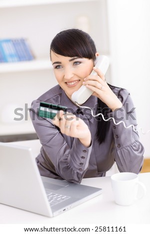 Smiling young women shopping online at home, holding credit card in hand and talking on phone.