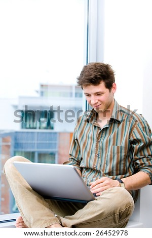 Young casual office worker sitting at office window working on laptop computer.