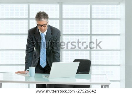 Worried businessman leaning on office desk with laptop computer on it,  looking down.