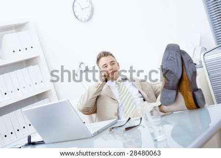 Satisfied businessman sitting by desk at office, feet on table, talking on mobile phone.