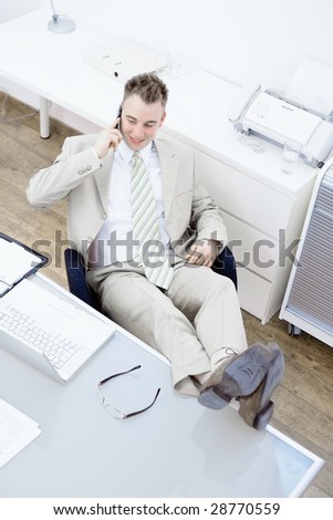 Satisfied businessman sitting by desk at office, feet on table, talking on mobile phone. High-angle view.