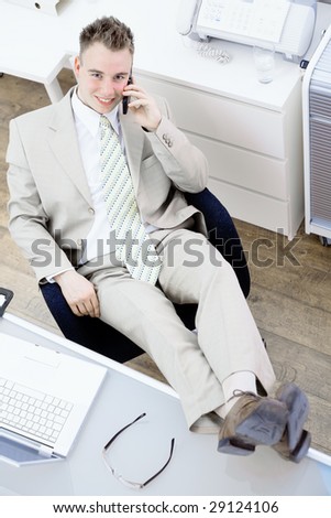 Satisfied businessman sitting by desk at office, feet on table, talking on mobile phone, smiling. High-angle view.
