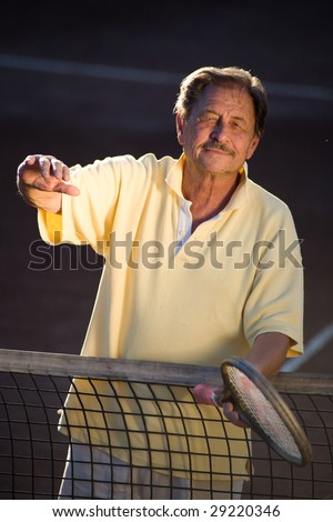 Active senior man in his 70s is offering his hand on the tennis court with tennis racket in other hand. Outdoor, sunlight.
