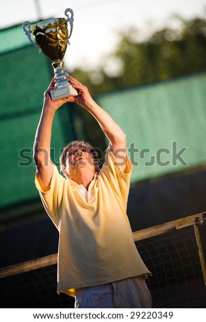 Active senior man in his 70s is posing on the tennis court with cup in hands. Outdoor, sunlight.