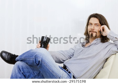 Relaxed man talking on mobile phone, sitting on a couch and holding a coffee cup in hand.