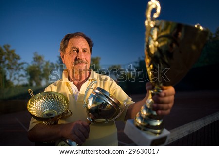 Active senior man in his 70s is posing on the tennis court with cups in hands. Outdoor, sunlight.
