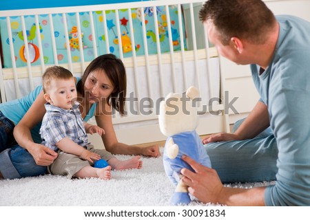 Portrait of happy family at home. Baby boy ( 1 year old ) and young parents father and mother sitting on floor and playing together at children\'s room, smiling.