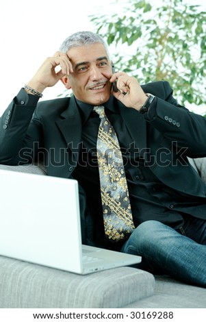 Gray haired mature businessman sitting on couch working on laptop computertalking on mobile phone, happy, smiling.
