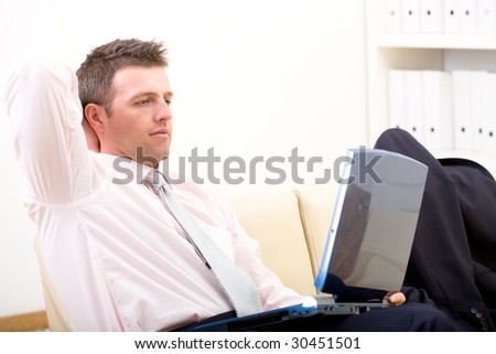 Relaxed businessman sitting at couch in office, using laptop computer, looking at screen.
