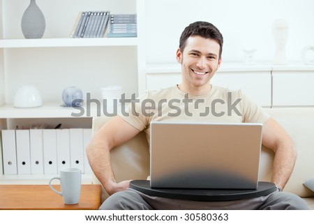 Happy man wearing beige t-shirt using laptop computer at home, sitting on couch, looking at camera, simling.
