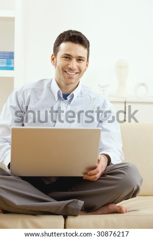 Happy young man sitting on couch and working on laptop computer at home, smiling.