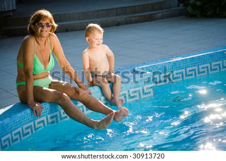 Grandmother and two years old granson enjoy water and sunshine on the poolside. Natural light, summer.