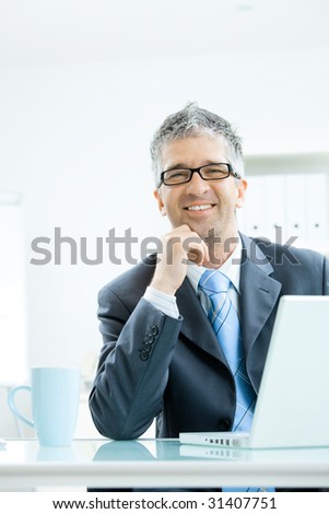 Businessman with grey hair, wearing grey suit and glasses thinking over laptop computer, sitting at office desk leaning on hand, smiling.