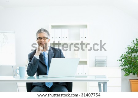 Businessman with grey hair, wearing grey suit and glasses thinking over laptop computer, sitting at desk in bright, modern office, leaning on hand, smiling.