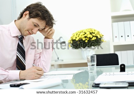 Young businessman wearing pink shirt, sitting at office desk and writing on paper, leaning on hand, thinking.