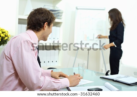 Young businessman sitting at desk, writing notes. Businesswoman standing in background, drawing chart on whiteboard. Selective focus on man.
