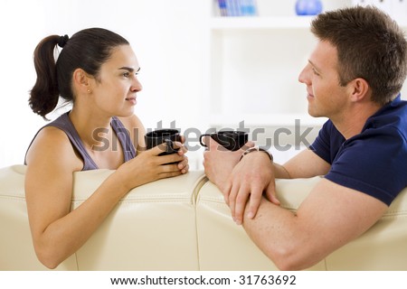 Adult couple sitting on sofa at home drinking coffe, talking, smiling.