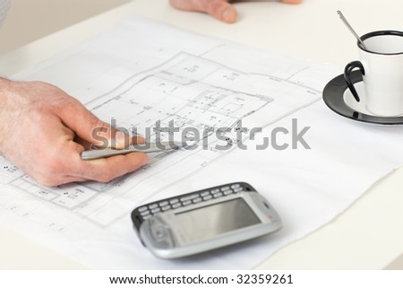 Architect\'s hand pointing with pen to floor plan on desk with mobile phone and coffee cup on it.
