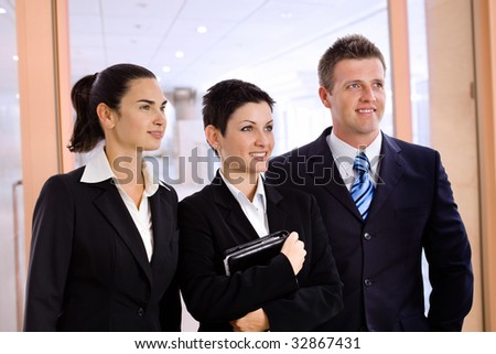 Happy successful young business people standing side by side at office lobby, smiling, looking away.