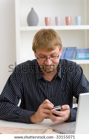 Man writing SMS at home, sitting at table with laptop and newspaper on it.