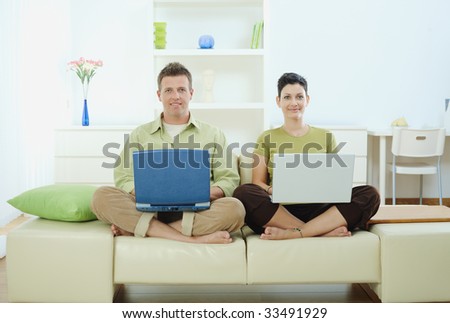 Happy young couple sitting on couch at home using laptop computer, smiling.