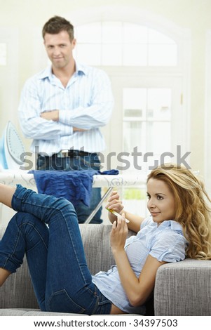 Relaxed young woman sitting on couch filing her nails, man watching with hands crossed behind ironing board.